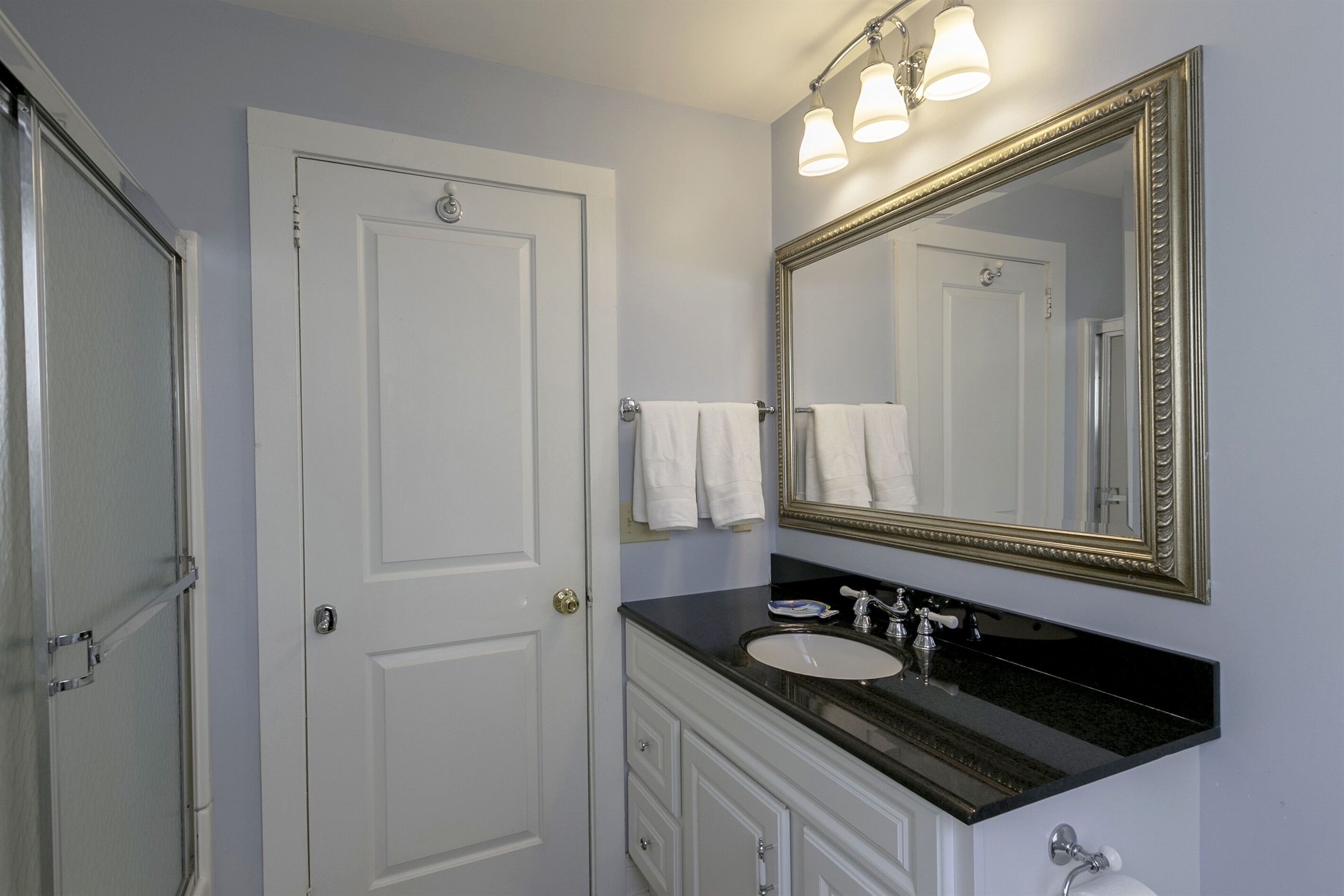 A bathroom with a white door, a large mirror, a black countertop, and a sink.