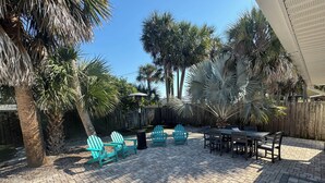 A patio area with brick flooring, outdoor furniture including chairs and a table, surrounded by palm trees and a wooden fence.