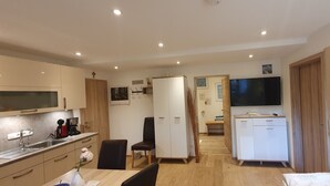 Eine moderne Küche mit Spüle, Herd und Schränken, ein Essbereich mit Tisch und Stühlen, ein an der Wand befestigter Fernseher und ein Flur, der zum Schlafzimmer führt.