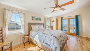 Ein Schlafzimmer mit einem Holzbett, Deckenventilator, einem Fenster mit Vorhängen, einem kleinen Tisch und Blick nach draußen.