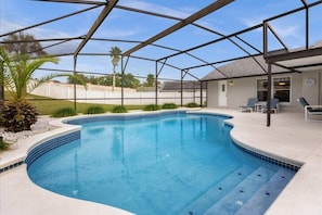 A swimming pool with a covered patio area, a house in the background, and a clear sky.