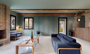 A modern living room with a brick fireplace, a blue sofa, a wooden coffee table, and a large window providing natural light. - Panorama Suite - Safran (Longiano)