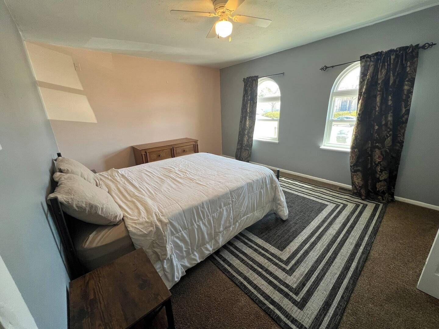 Une chambre à coucher avec un lit, une table de chevet, un ventilateur de plafond et une fenêtre avec des rideaux.
