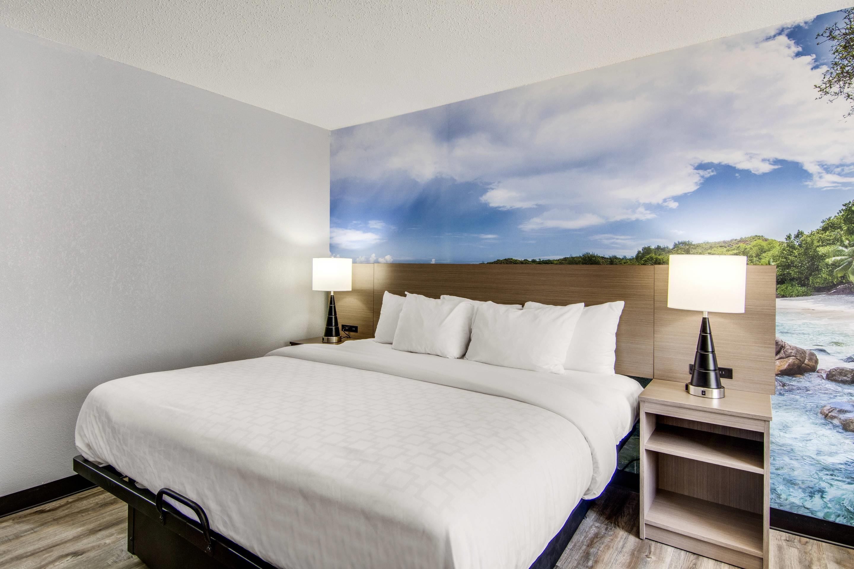 A hotel room with a large bed, two bedside tables with lamps, a headboard, and a mural of a landscape.