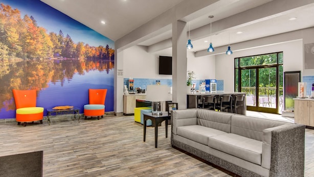 A modern hotel lobby with a large mural of a lake and autumn trees, a seating area with colorful chairs, a sofa, and a kitchen area with a large window.
