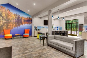 A modern hotel lobby with a large mural of a lake and autumn trees, a seating area with colorful chairs, a sofa, and a kitchen area with a large window. - Clarion Pointe on the lake Clarksville-South Hill West (Clarksville)