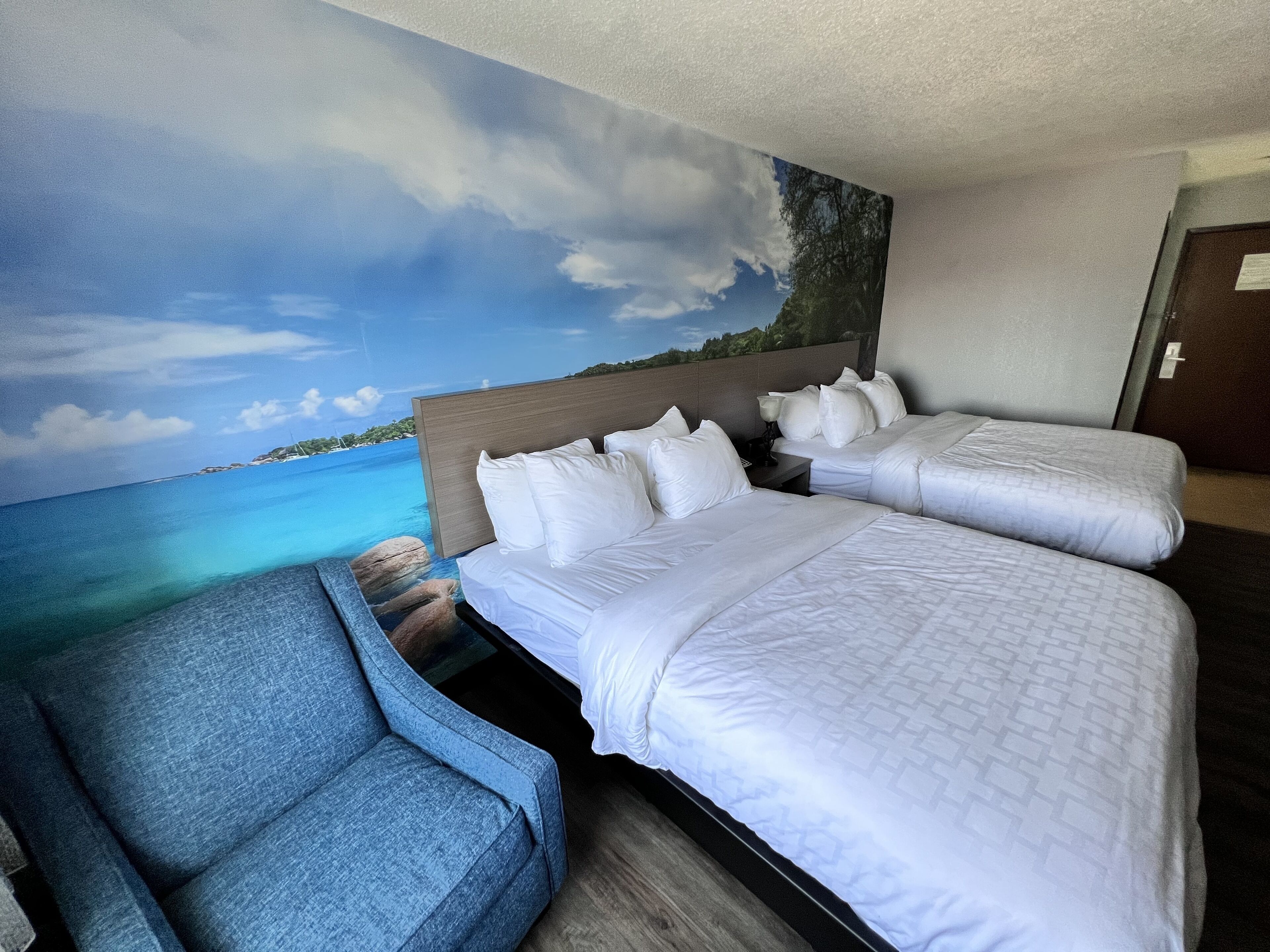 A hotel room with two beds, a blue armchair, and a mural of a beach scene.
