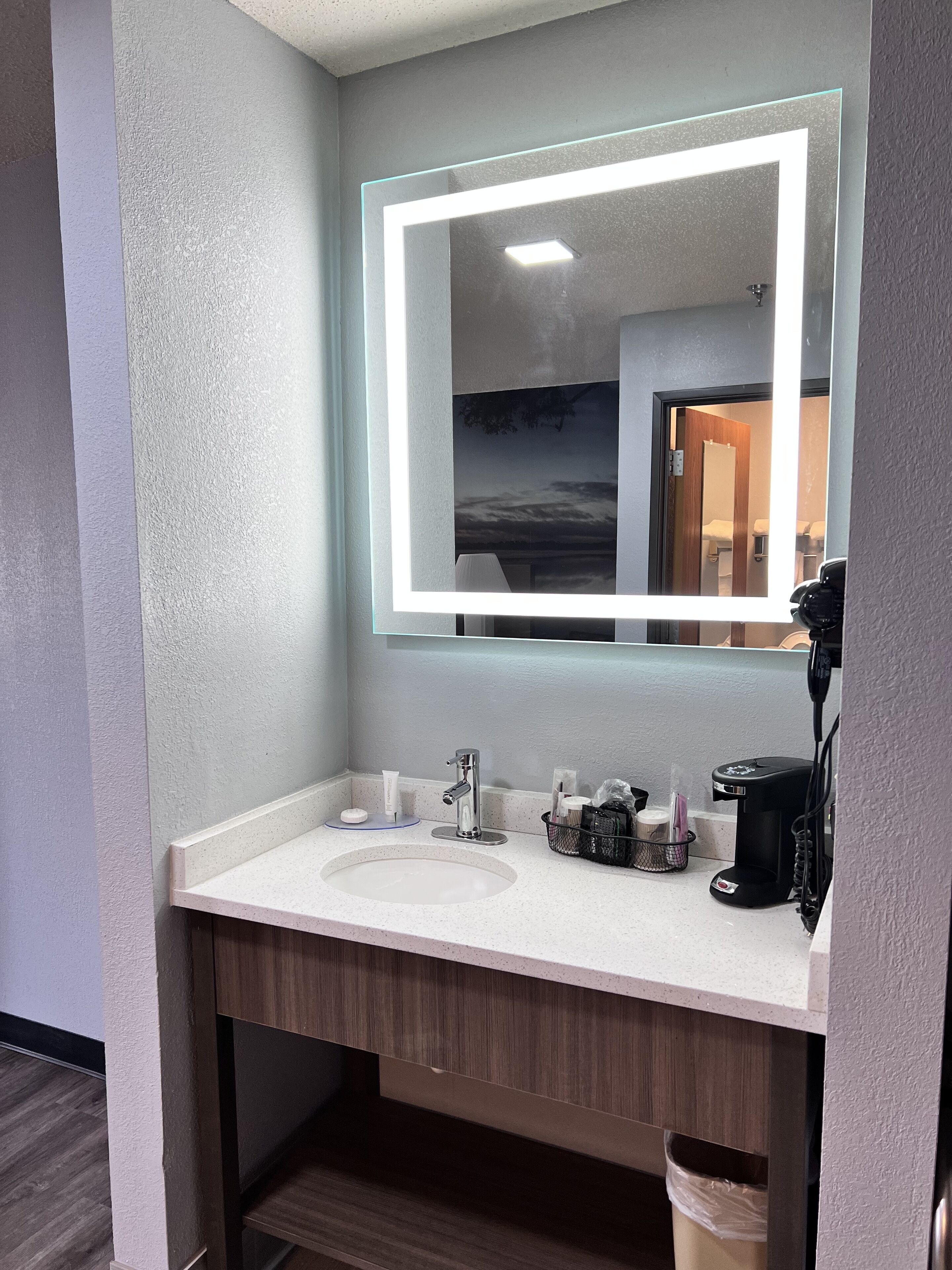 Une salle de bain moderne avec un lavabo blanc, un grand miroir doté d’un éclairage à DEL et une machine à café posée sur le comptoir.