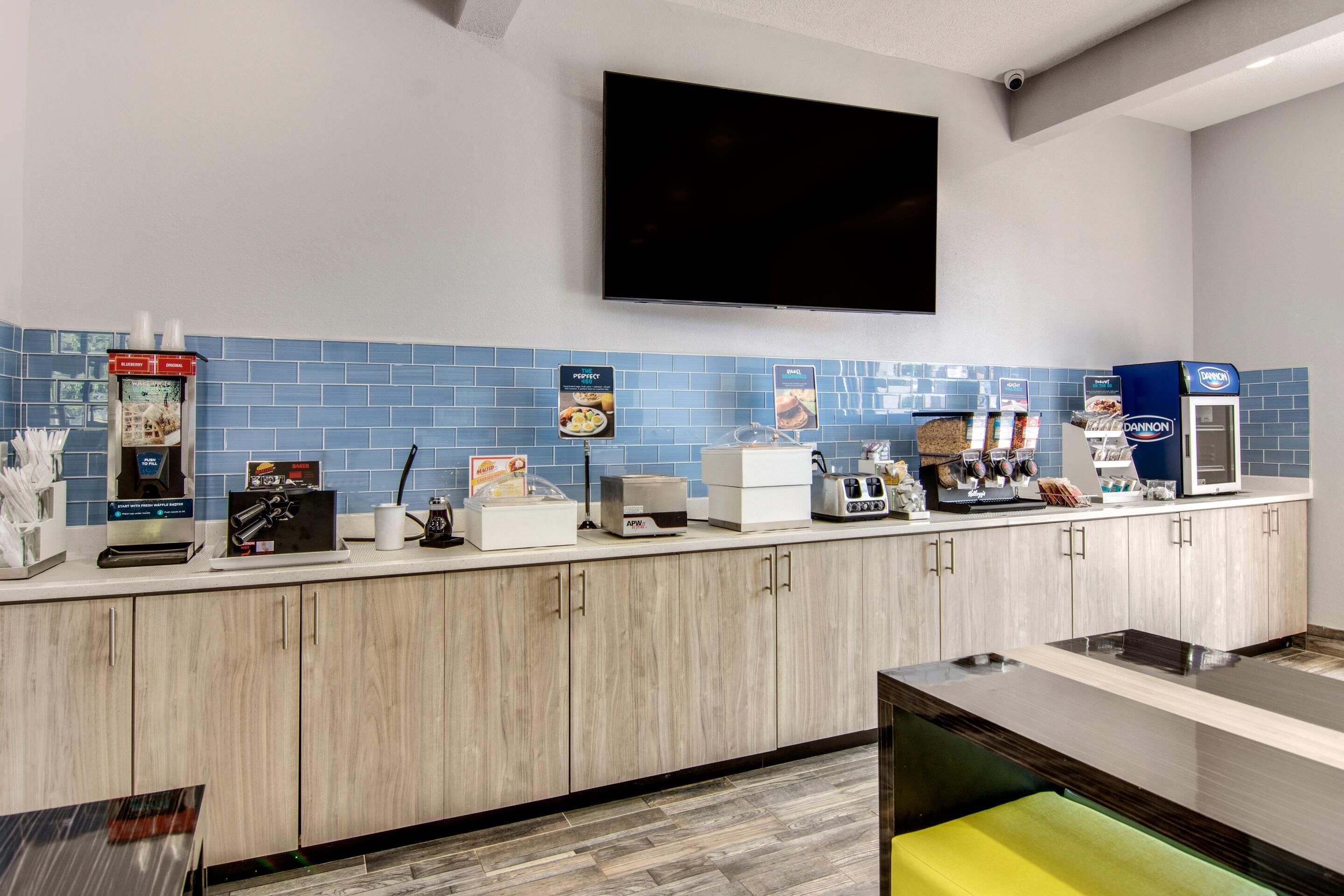 A modern kitchen area with a flat-screen TV mounted on the wall, a coffee machine, a waffle maker, and a bread machine.