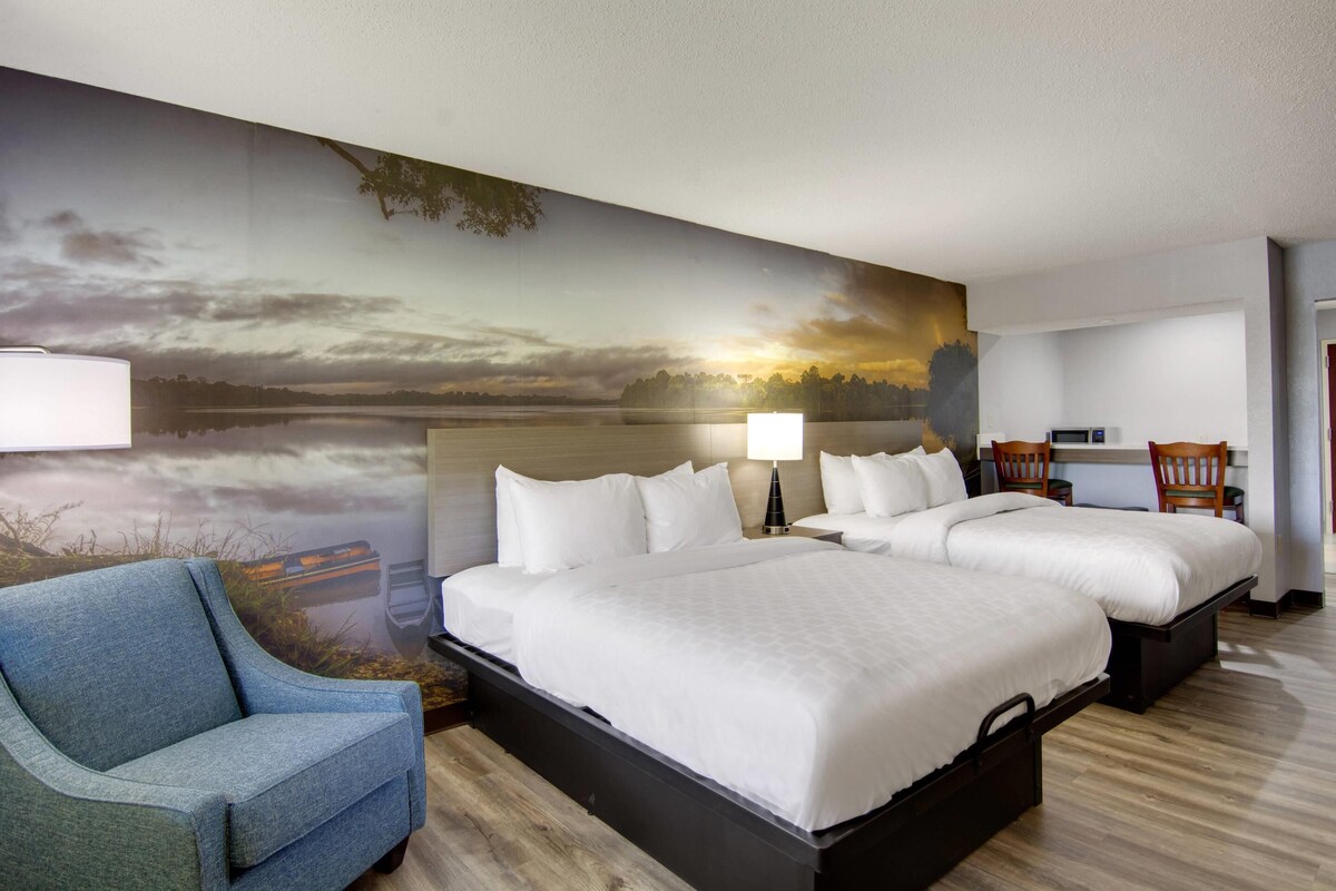 A hotel room with two beds, a blue armchair, a desk with a chair, and a large scenic wall mural.