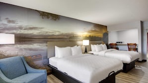 A hotel room with two beds, a blue armchair, a desk with a chair, and a large scenic wall mural.