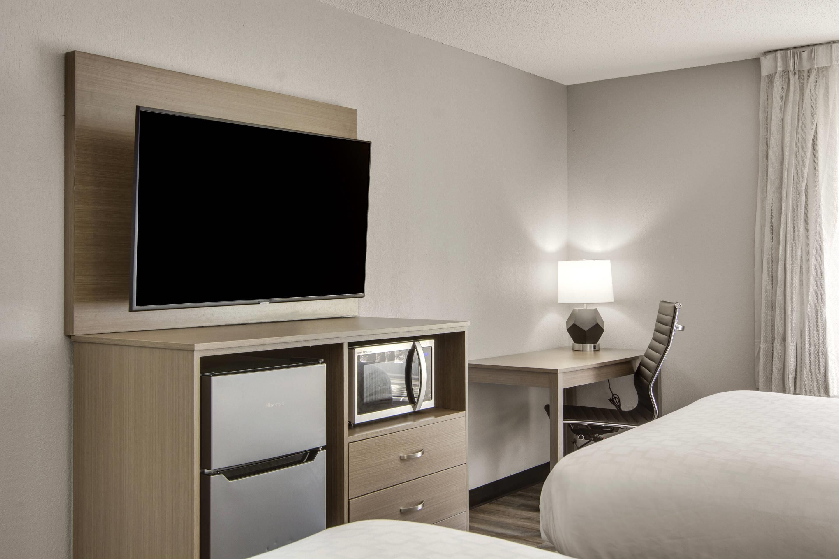 A hotel room with a bed, a desk with a lamp, a television, a minibar, and a chair.