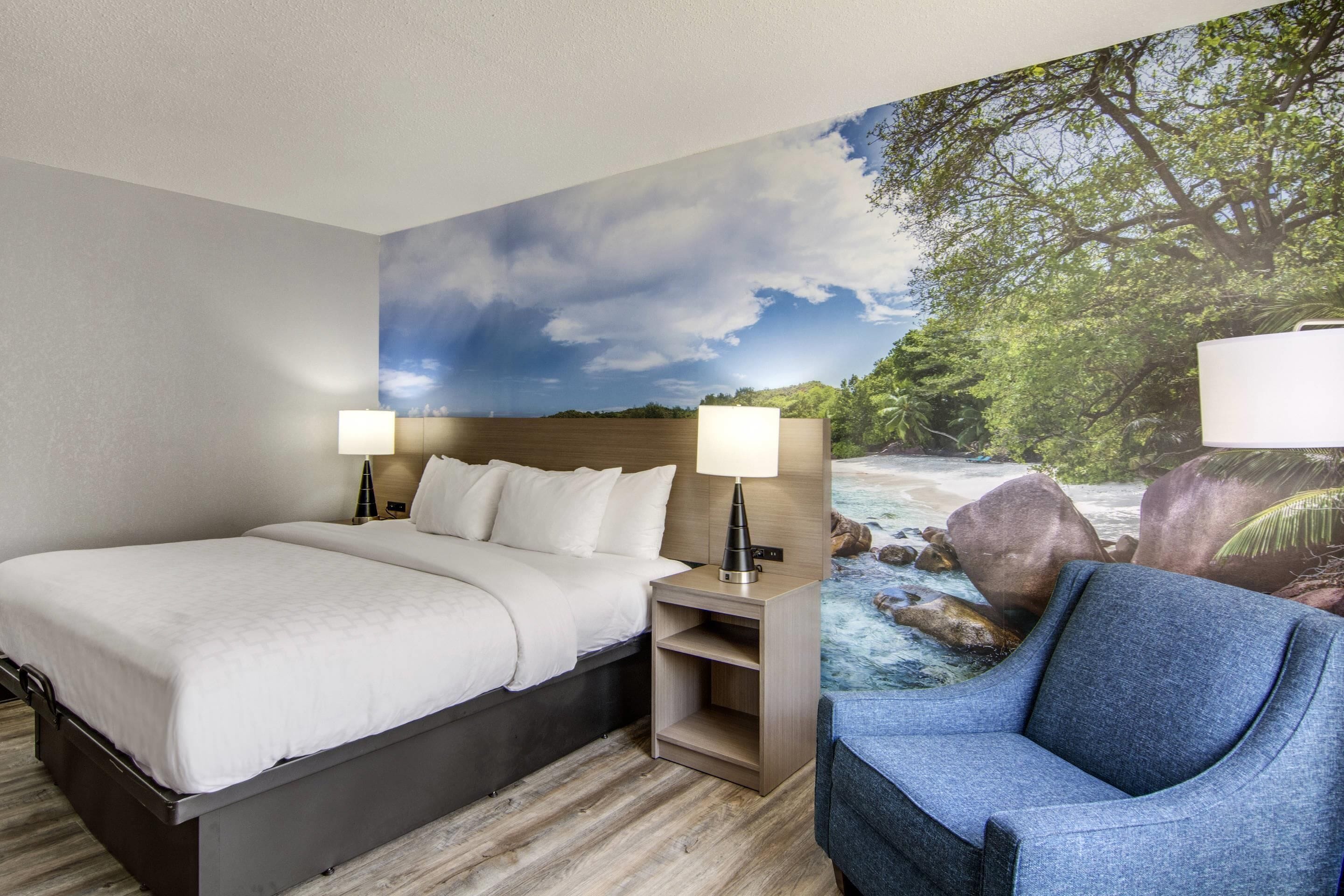 Ein Hotelzimmer mit Bett, zwei Nachttischlampen, einem Sessel und einem Wandgemälde von einem Fluss und Bäumen.