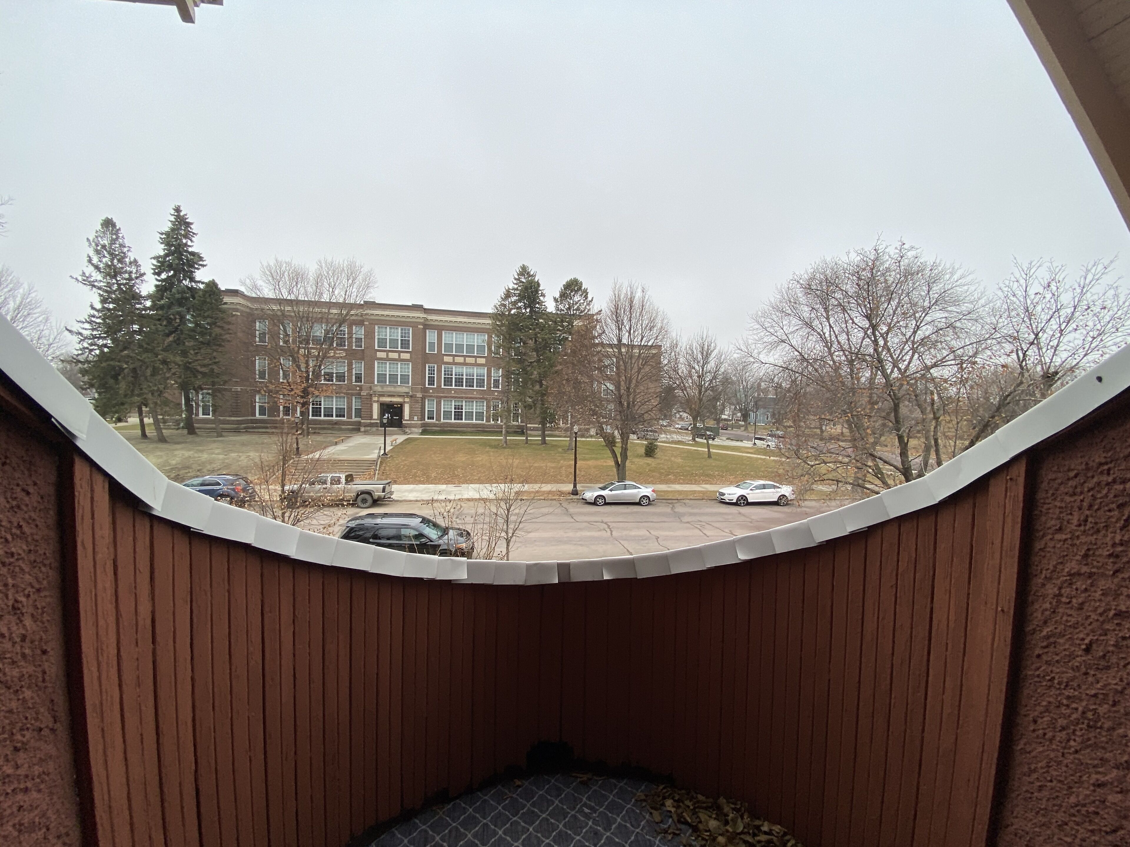 A view from a window with a curved wooden frame, overlooking a parking lot with several cars and a multi-story building with a brick facade.