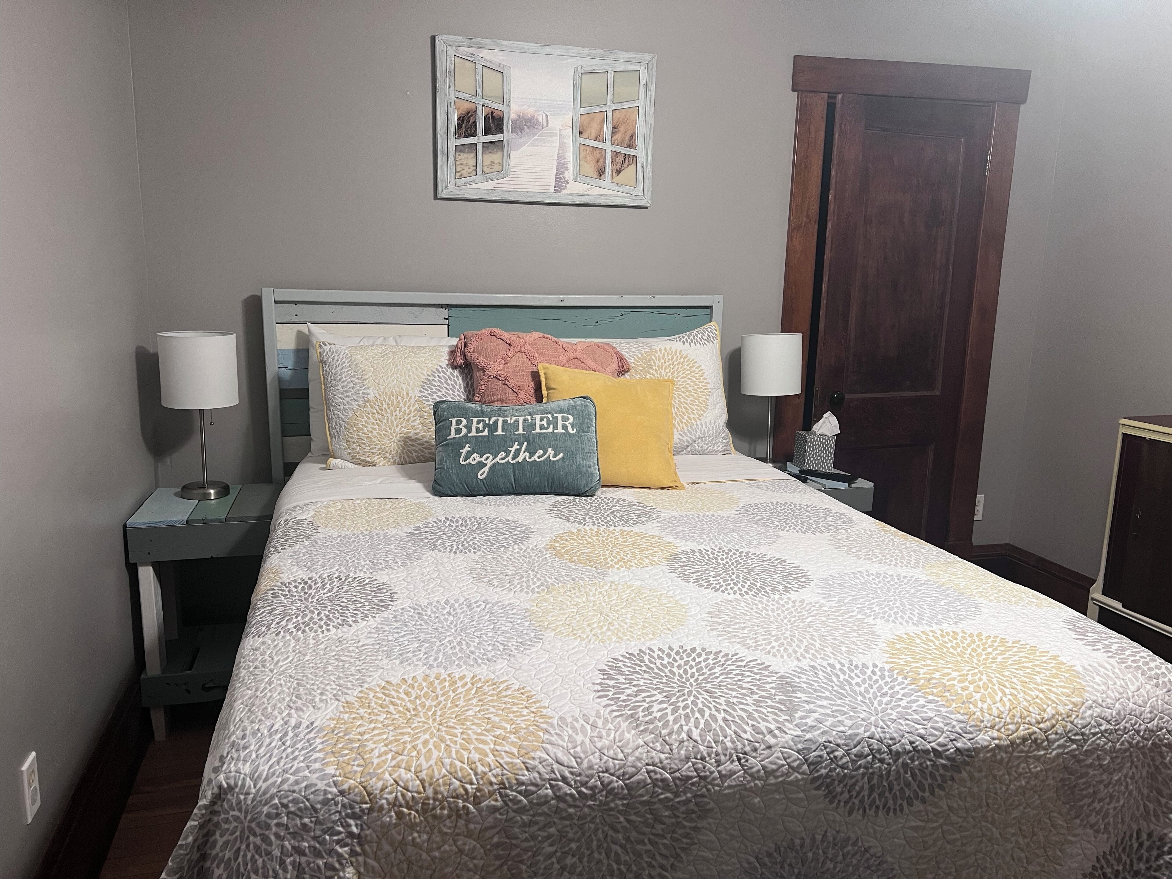 A neatly made bed with a patterned bedspread, a pillow with the text "BETTER together," and a decorative wall piece above it.