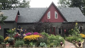 Une maison à l’extérieur d’un gris foncé et au toit rouge, entourée d’un jardin bien entretenu où poussent diverses fleurs et plantes.