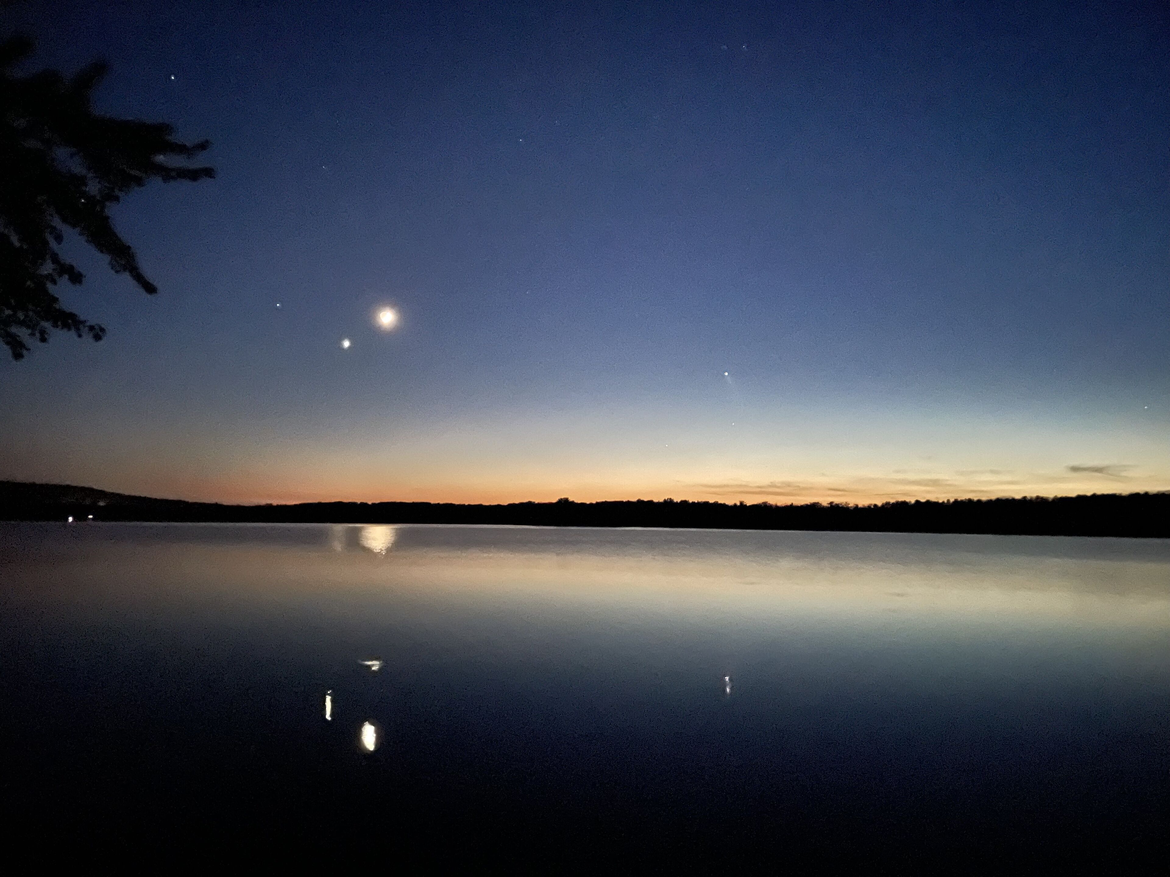 Um lago calmo ao entardecer com céu limpo, refletindo a lua e as estrelas.
