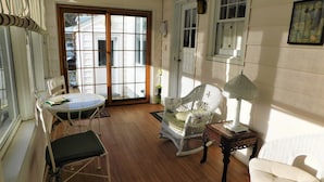 Quarto com piso de madeira, uma mesa branca com um livro, uma cadeira de vime, um poltrona branca e um abajur.