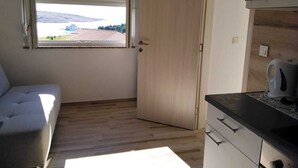 Een kleine kamer met een slaapbank, een houten deur, een raam met jaloezieën en een kitchenette.