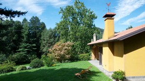 Un bâtiment jaune, doté d’une cheminée et d’un girouette, entouré d’un jardin bien entretenu avec une pelouse, des arbres et une chaise en bois.