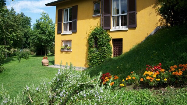 Une maison jaune aux volets bruns, entourée d’un jardin bien entretenu où poussent différentes plantes et fleurs.