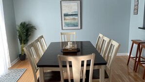 Una sala da pranzo con un tavolo in legno scuro e sedie bianche, una ventola a soffitto, un quadro appeso al muro e una pianta in vaso.