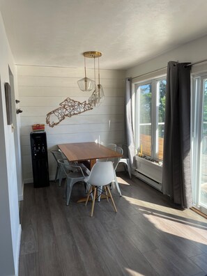 Un comedor con mesa de madera, sillas blancas, lámparas colgantes, dispensador de agua y una ventana con cortinas.