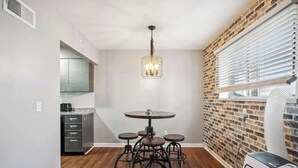 A modern kitchen with a round dining table and stools, a hanging light fixture, and a brick accent wall. - Fairlane #2 | Uncaged (Lansing)