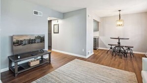 A modern living room with a flat-screen TV, dark wood flooring, a dining area with a round table and chairs, and a chandelier. - Fairlane #2 | Uncaged (Lansing)