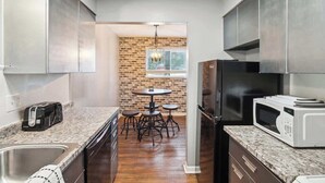 A modern kitchen with granite countertops, stainless steel appliances, a brick accent wall, and a dining area with a round table and chairs. - Fairlane #2 | Uncaged (Lansing)