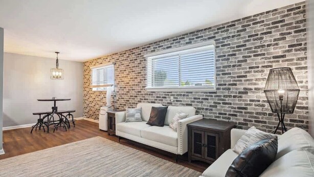 A living room with a brick wall, a white sofa, a dining table with chairs, and a lamp. - Fairlane #2 | Uncaged (Lansing)