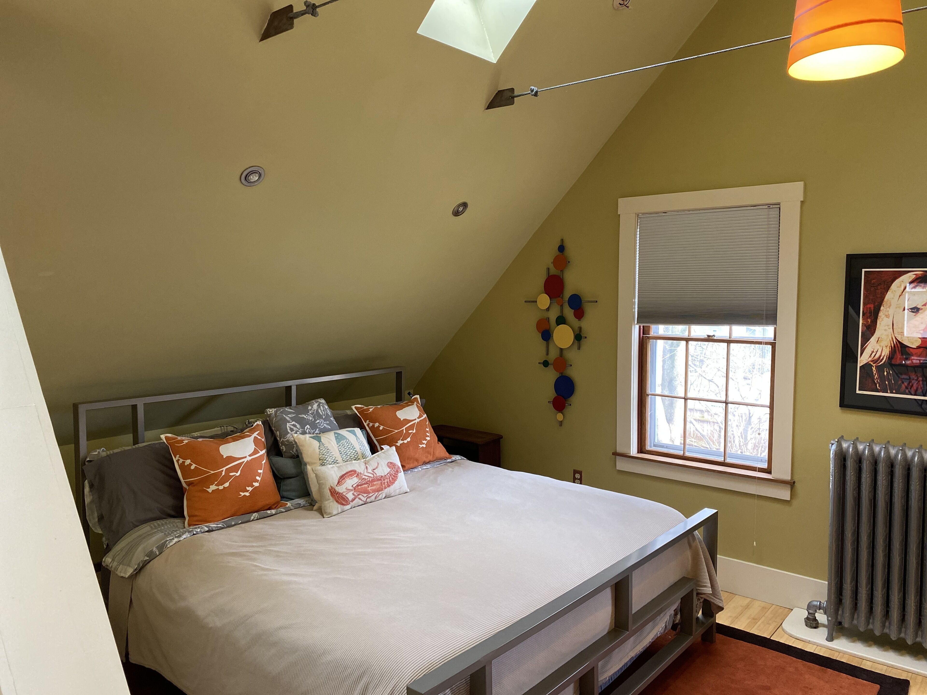 Une chambre avec un plafond en pente, un lit avec de nombreux oreillers, une fenêtre avec des stores, un radiateur et un luminaire fixé au mur.