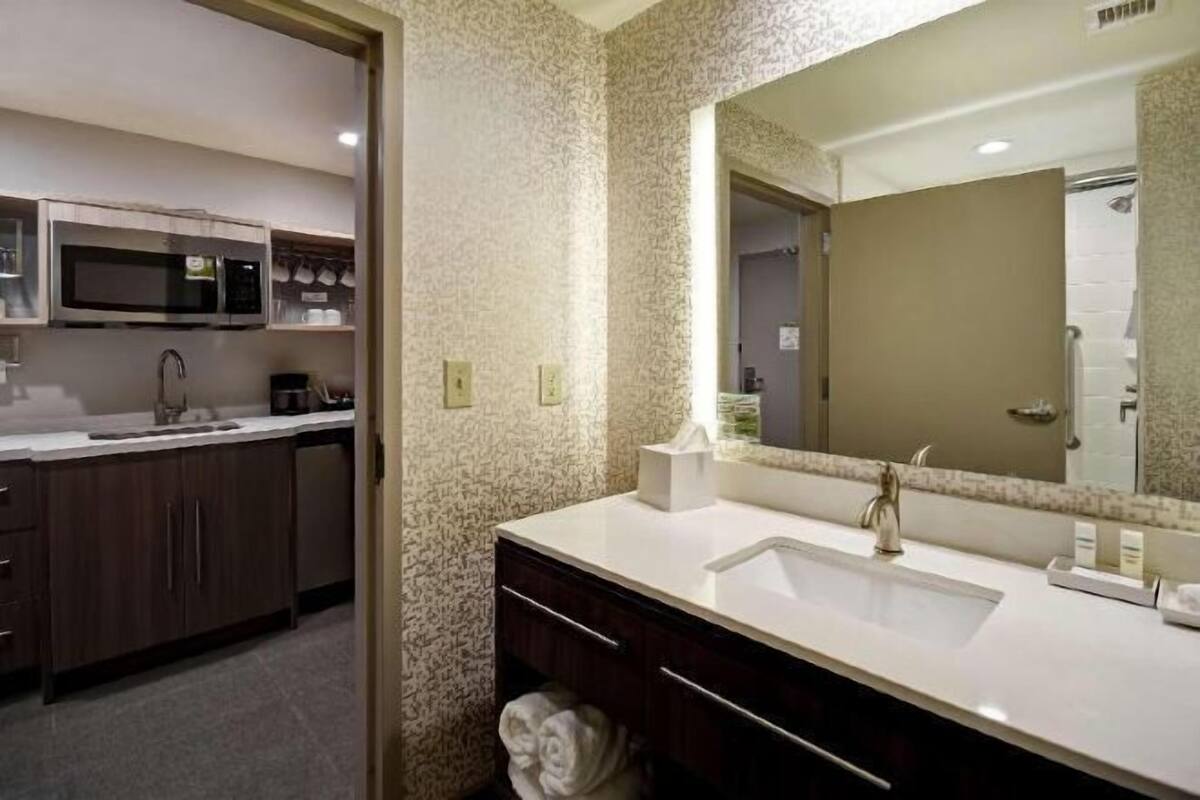 A hotel bathroom with a double sink, a large mirror, and a microwave in the adjacent kitchenette.