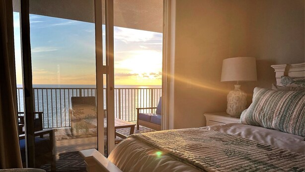 A bedroom with a bed, a nightstand, a lamp, and a view of the ocean through a sliding door.