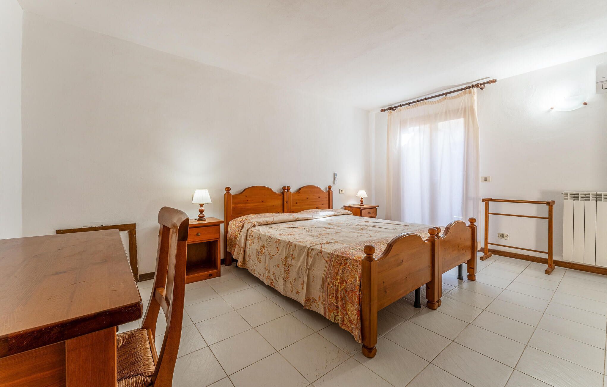 A bedroom with a wooden bed, bedside tables with lamps, a wooden desk, and a radiator.