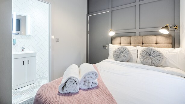 A neatly made bed with two rolled towels, a headboard with a light fixture, and a bathroom with a white sink and mirror. - Swansea's Hub (Swansea)