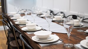 A dining table set with white plates, bowls, and clear wine glasses, arranged in a row with black chairs. - Swansea's Hub (Swansea)