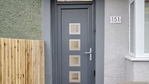 A grey door with four square windows, a metal handle, and a mailbox below. A wooden fence is visible to the left, and a house number '151' is on the wall to the right. - Swansea's Hub (Swansea)
