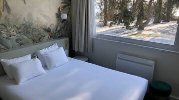 A hotel room with a bed, a nightstand, a window with a view of trees and snow, and a wall mural.