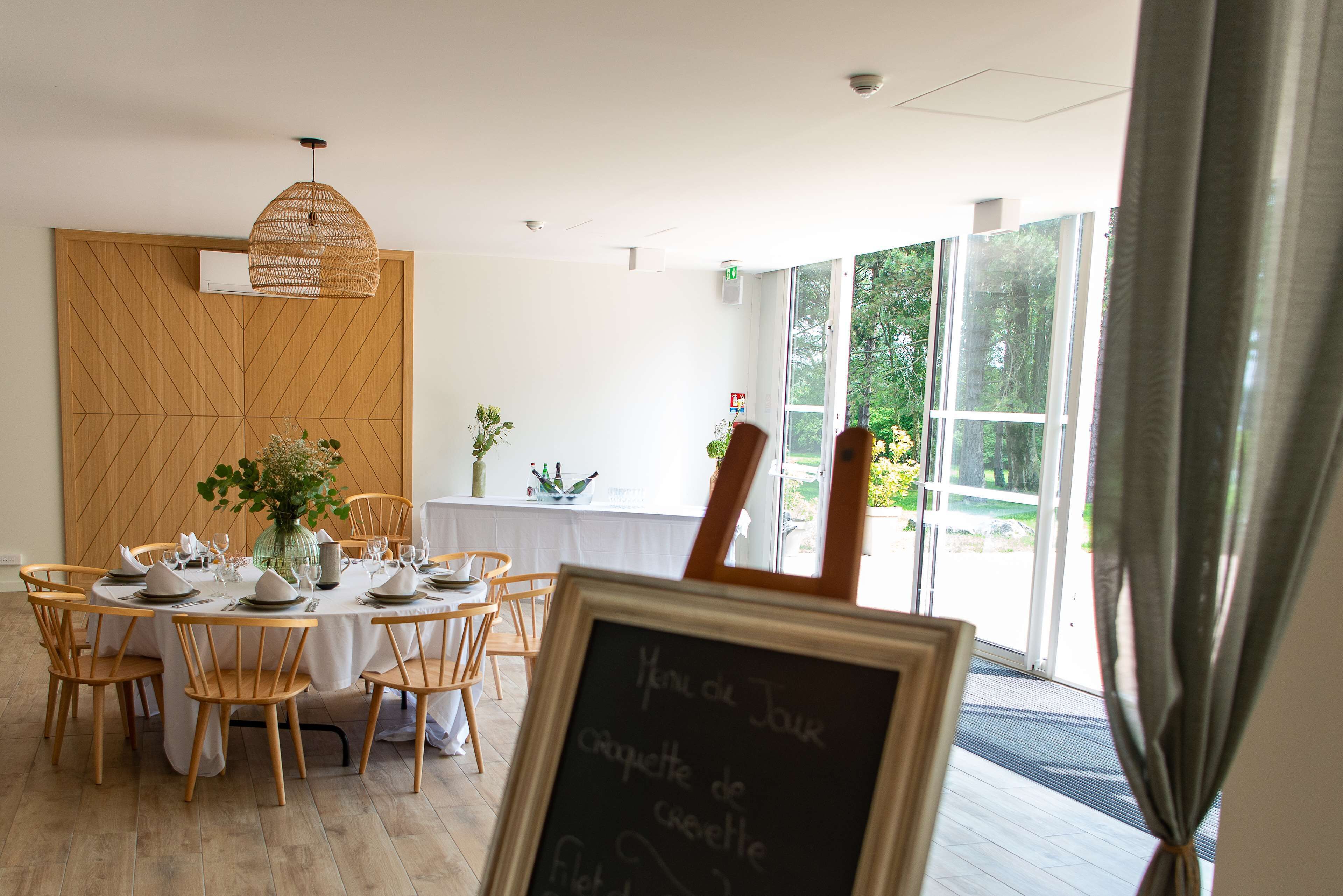 A dining area with a round table set for six, wooden chairs, a hanging light fixture, and a chalkboard sign.