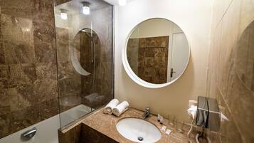 Une salle de bain moderne avec une douche à parois de verre, un miroir rond, un lavabo avec des articles de toilette et un mur recouvert de carreaux de marbre.