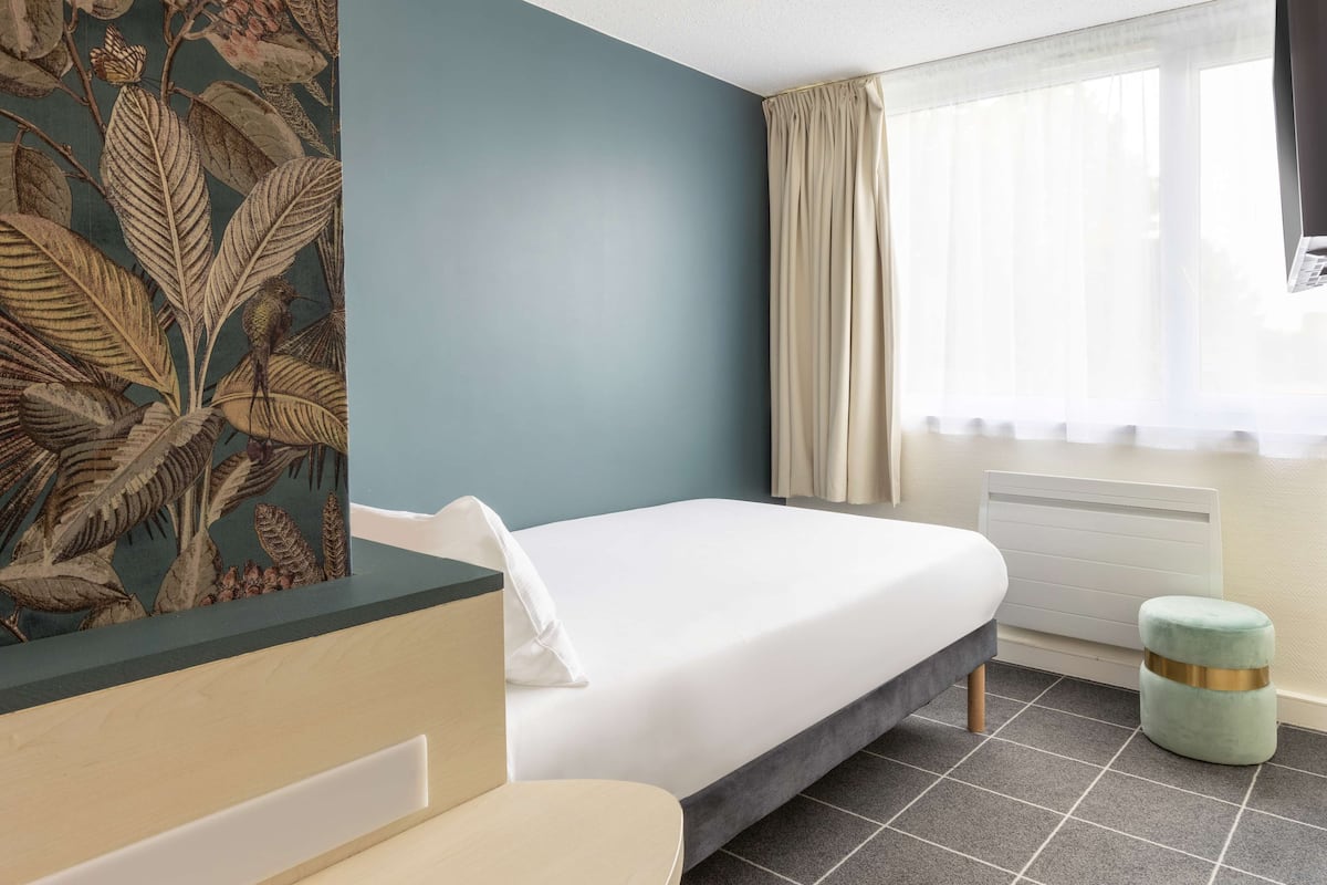 Une chambre d’hôtel avec un lit, une petite table, une chaise, une fenêtre avec des rideaux et un radiateur.