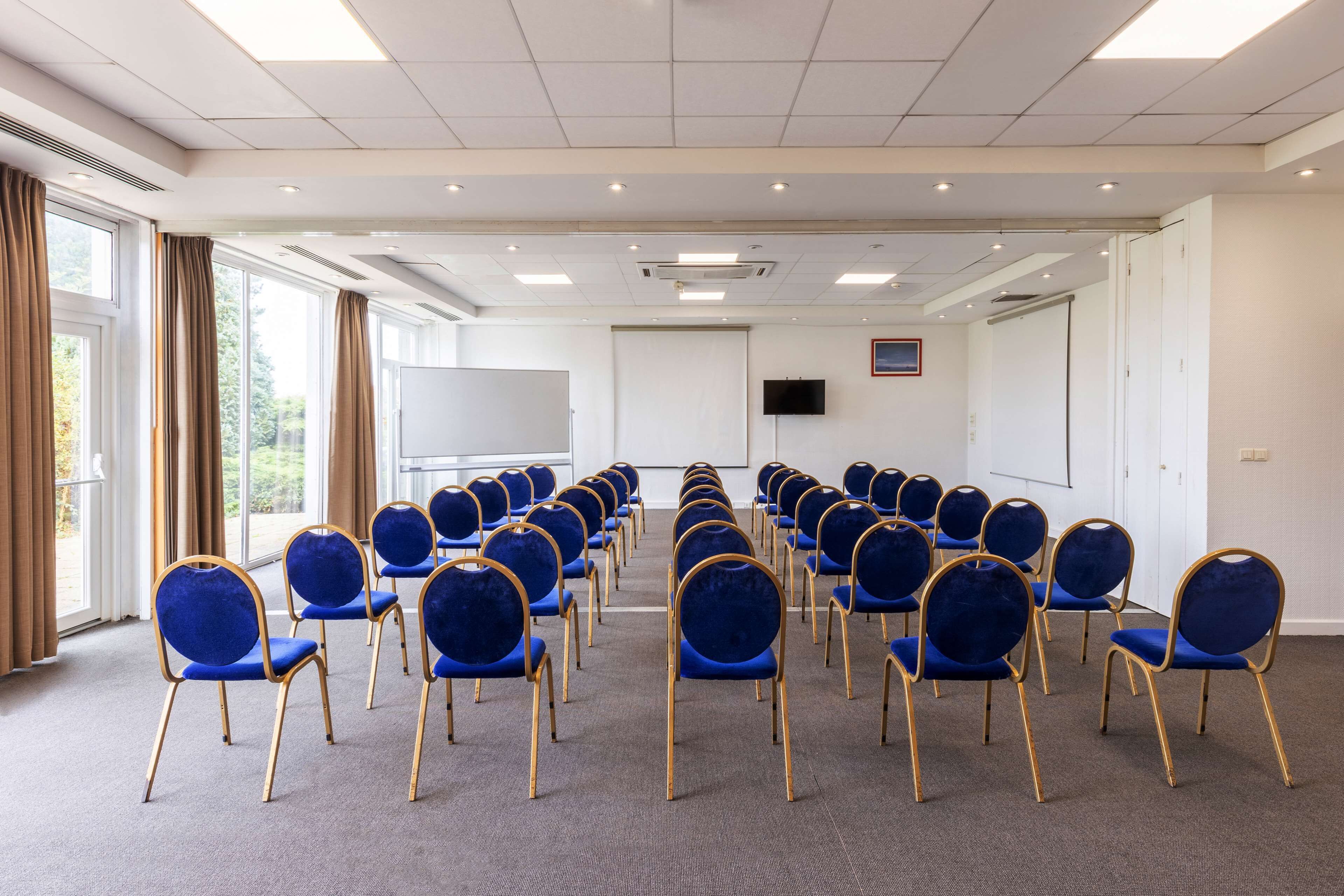 Une salle de conférence avec des rangées de chaises bleues, un tableau blanc et un écran de projection.