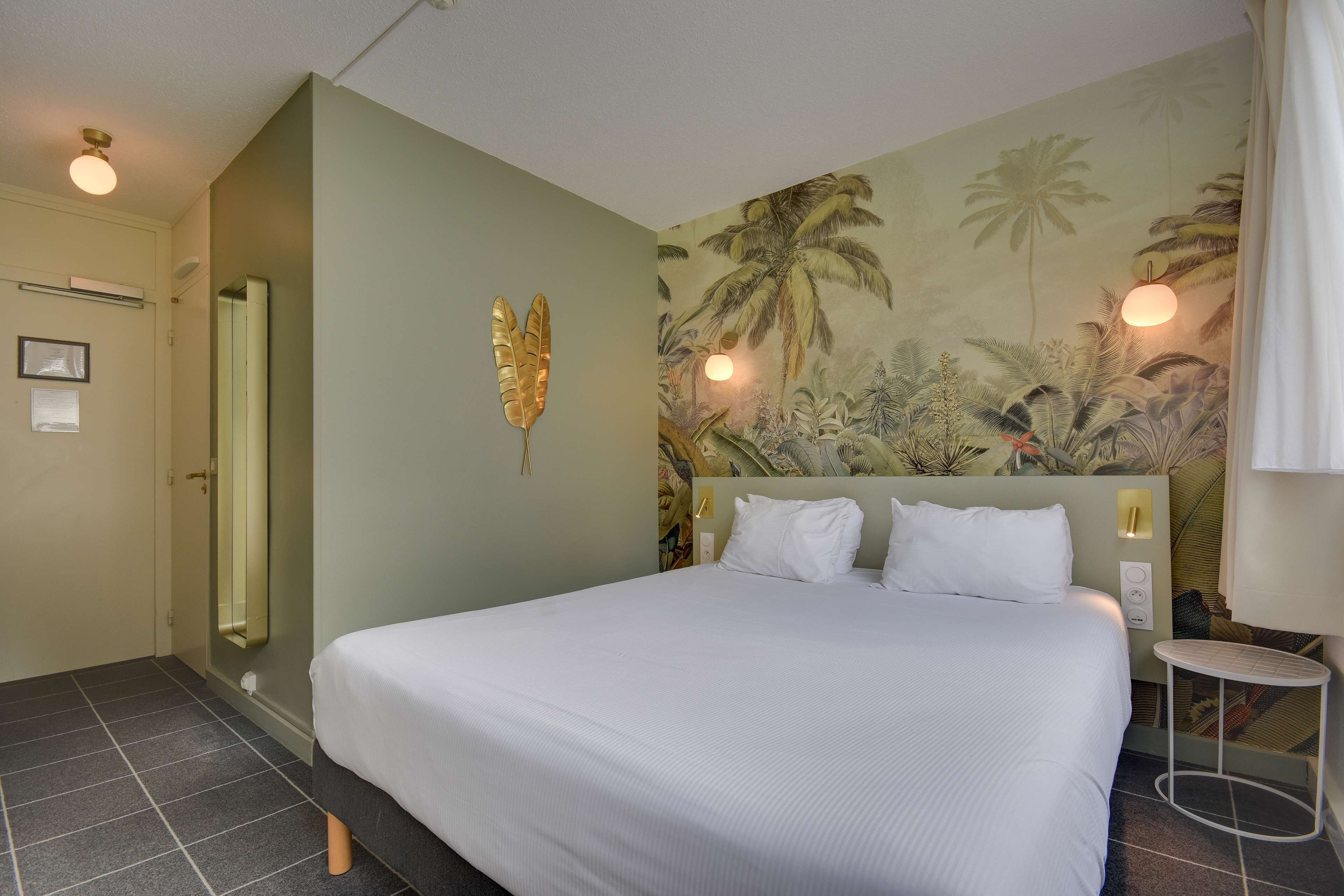 A neatly made bed with white linens, a wall-mounted mirror, a small bedside table, and a tropical-themed mural on the wall.