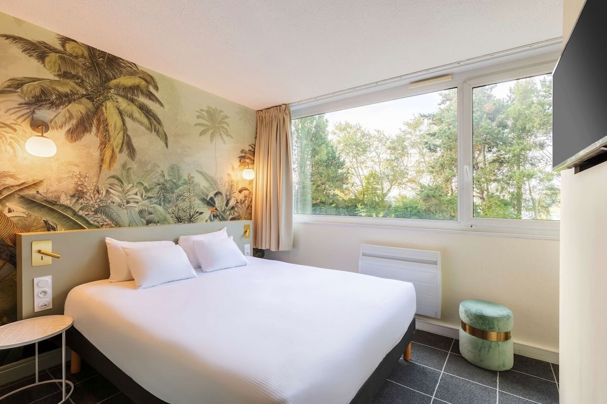 Une chambre d’hôtel comprenant un lit, une petite table, une chaise, un radiateur, une fenêtre donnant sur des arbres et un mur orné d’une fresque aux motifs tropicaux.