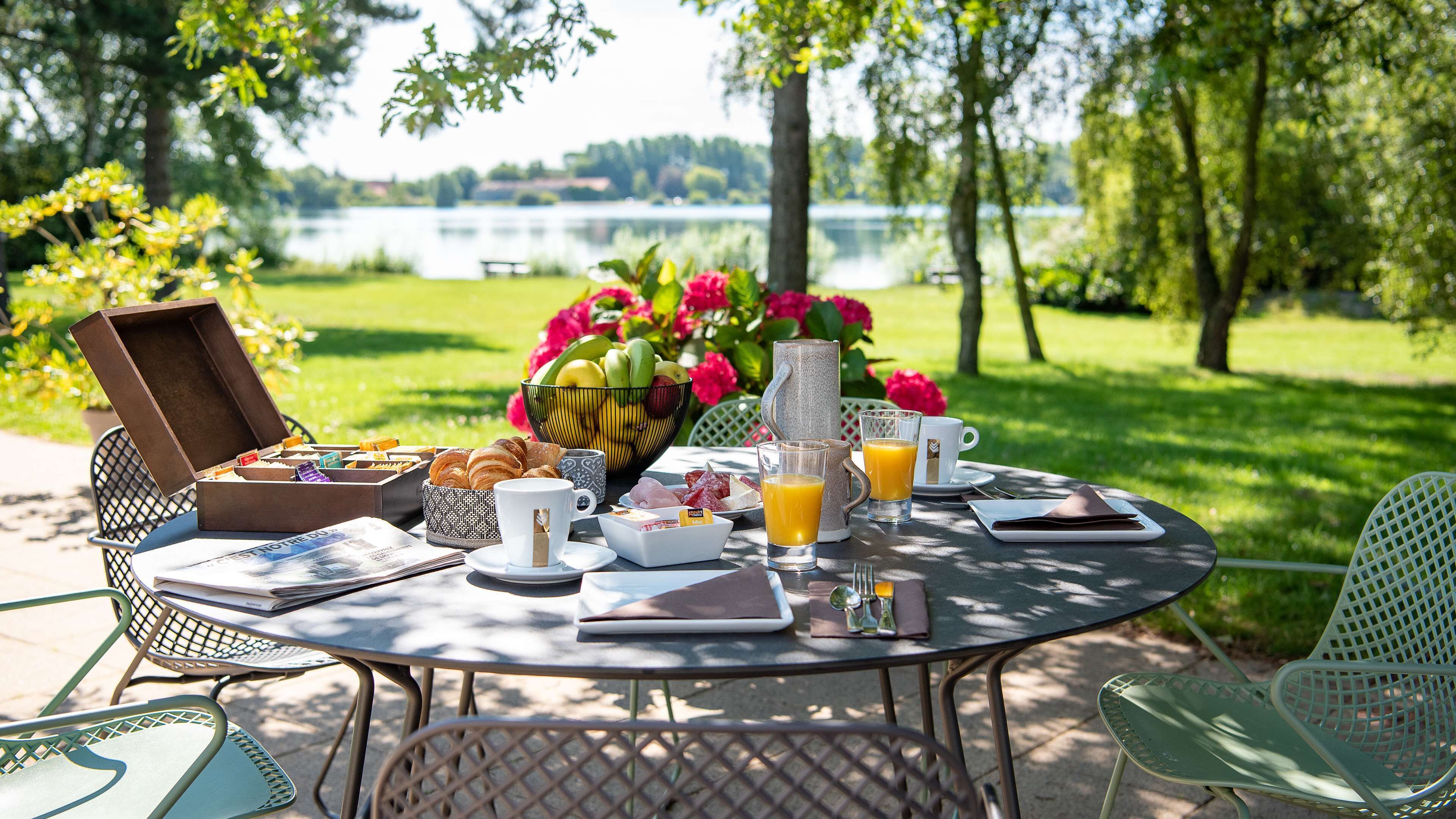 Une table dressée pour le petit-déjeuner, avec des croissants, des fruits et du jus, placée à l’extérieur, avec des chaises, et offrant une vue sur un lac.