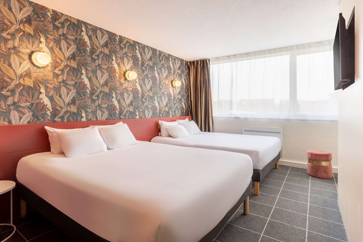 Une chambre d’hôtel avec deux lits, une tête de lit rouge, un mur à motifs, un téléviseur fixé au mur et une fenêtre avec des rideaux.
