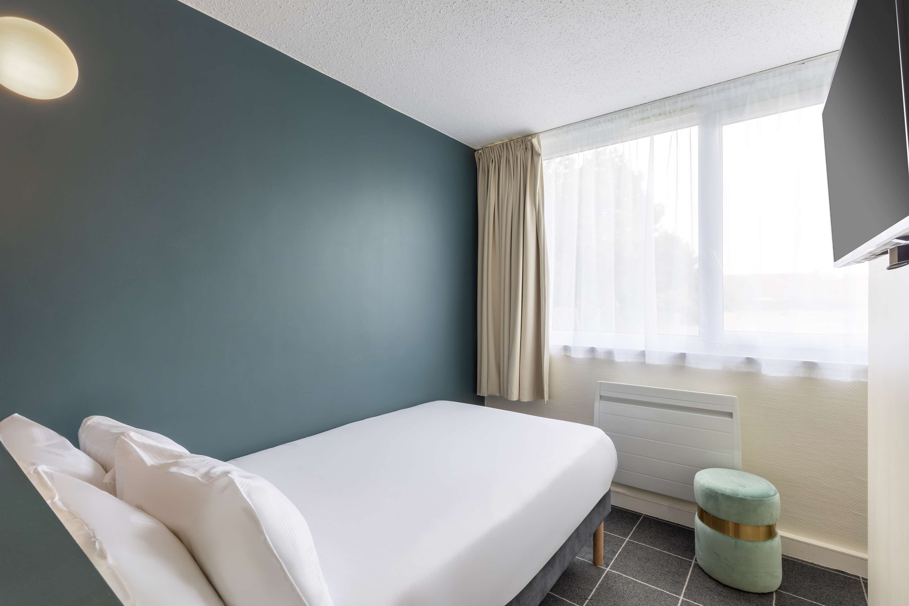 Un lit simple dans une chambre aux murs de couleur turquoise, une fenêtre avec des rideaux légers, un petit tabouret et un radiateur blanc.