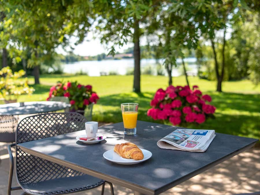 Une table à manger en plein air avec des chaises, un croissant, un verre de jus d’orange et un journal.