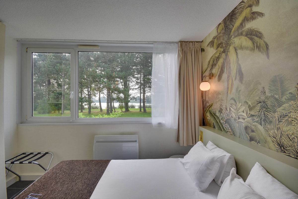 Une chambre d’hôtel avec un lit, une fenêtre donnant sur des arbres, un radiateur et un mur orné d’une fresque aux motifs tropicaux.